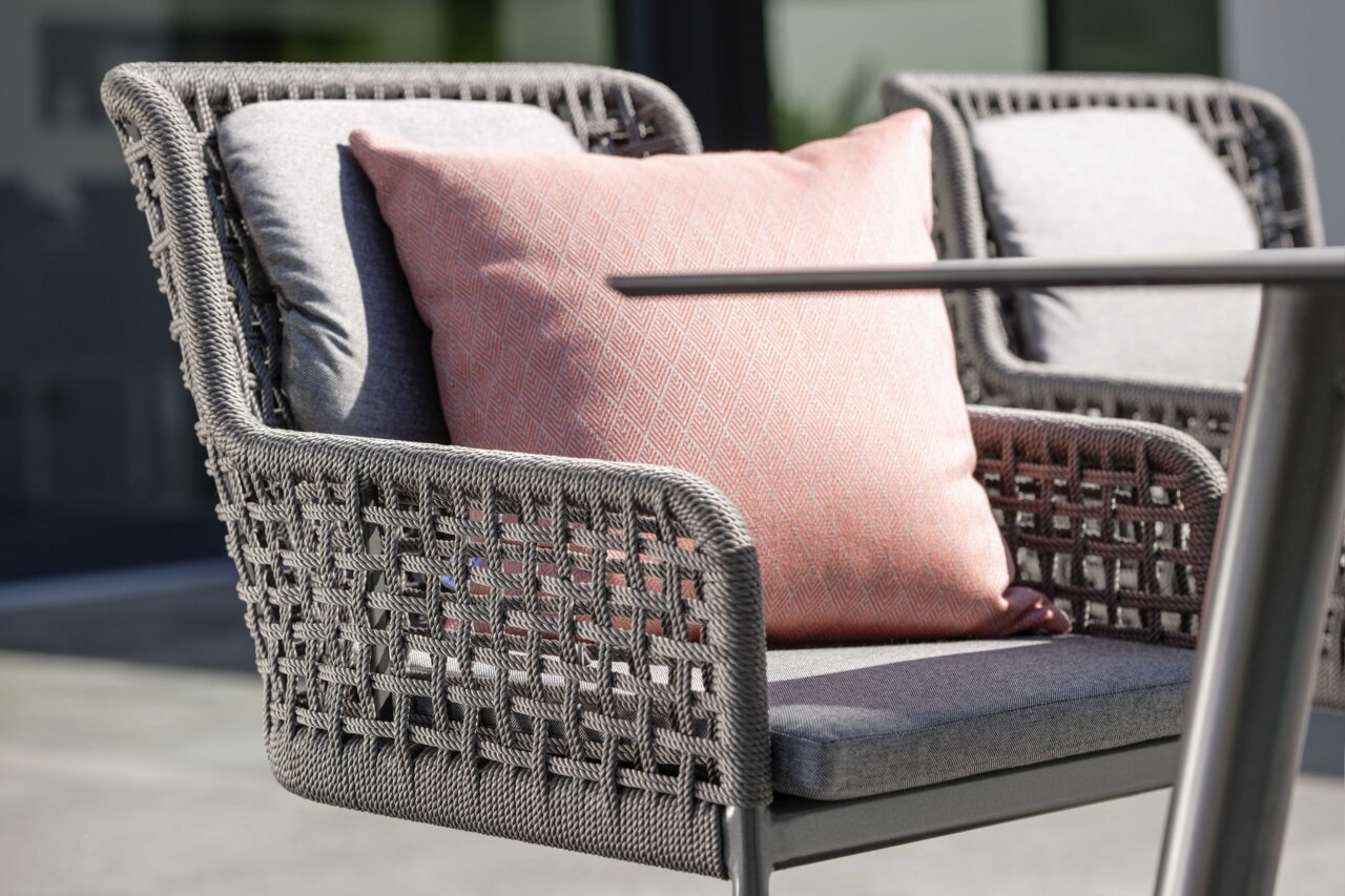 Nahaufnahme eines modernen Freischwingersessels mit grauem Geflecht und einem rosa Kissen, seitliche Perspektive.