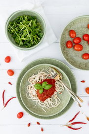 Draufsicht auf ein gedecktes Tisch-Arrangement mit grünen Keramikschalen: Pasta mit Tomatensauce und Basilikum, Rucola in einer Schüssel sowie Kirschtomaten und Chilischoten auf weißem Holztisch.