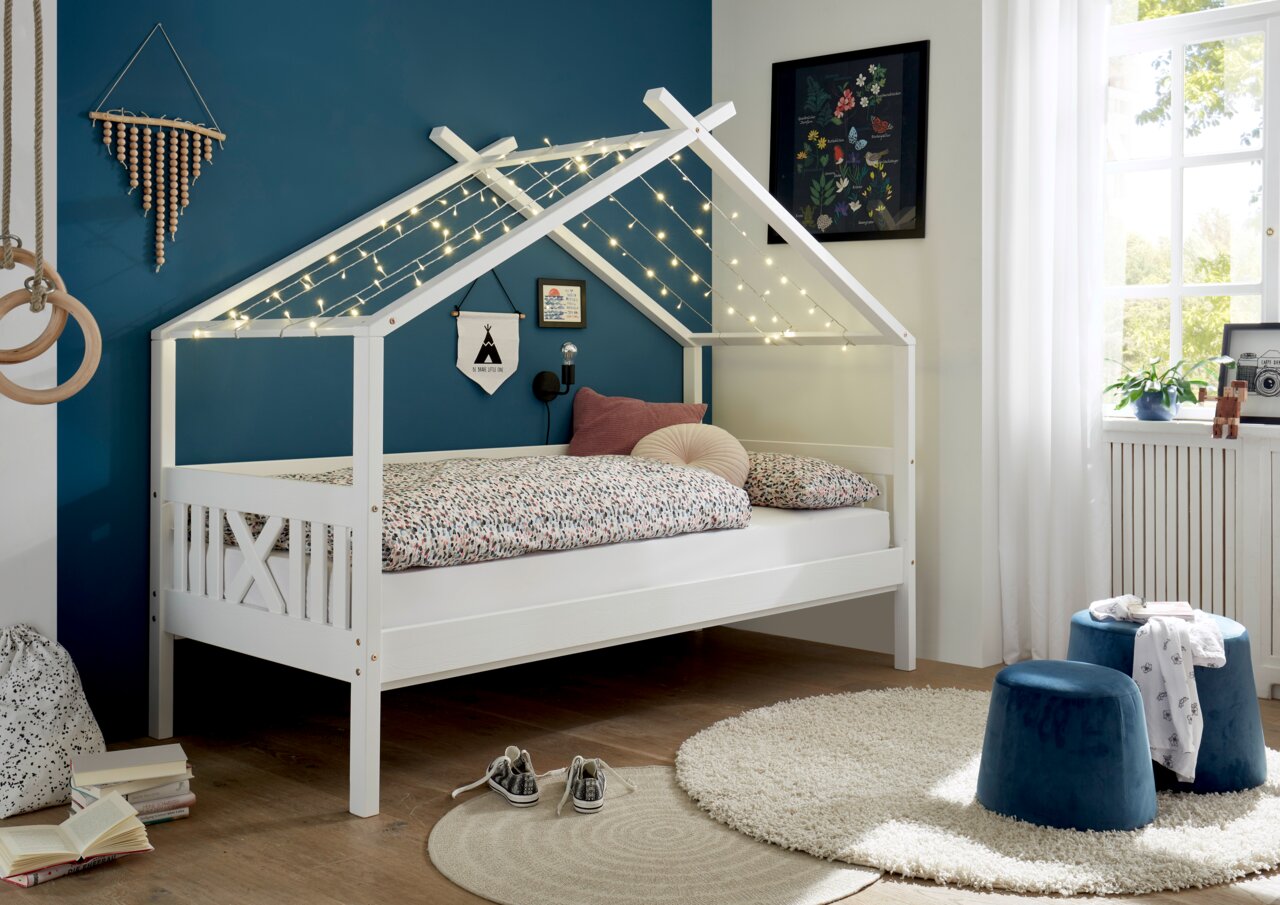 Weißes Hausbett mit Lichterkette, seitliche Perspektive, dekoriert mit bunter Bettwäsche und Kissen, in einem gemütlichen Kinderzimmer.