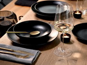 Schwarz-mattes Geschirr-Set mit tiefem Teller und Schale, goldfarbenem Besteck und Weinglas auf Holztisch mit Teelichtern, fotografiert in schräger Nahaufnahme von oben (3/4-Perspektive).