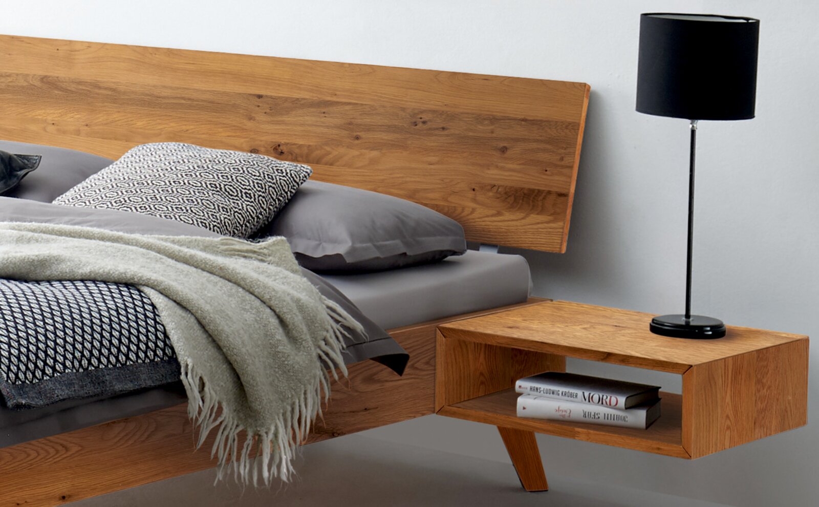 Holzbettgestell mit Kopfteil aus Holz, grauer Bettwäsche und Nachttisch mit Lampe, seitliche Perspektive