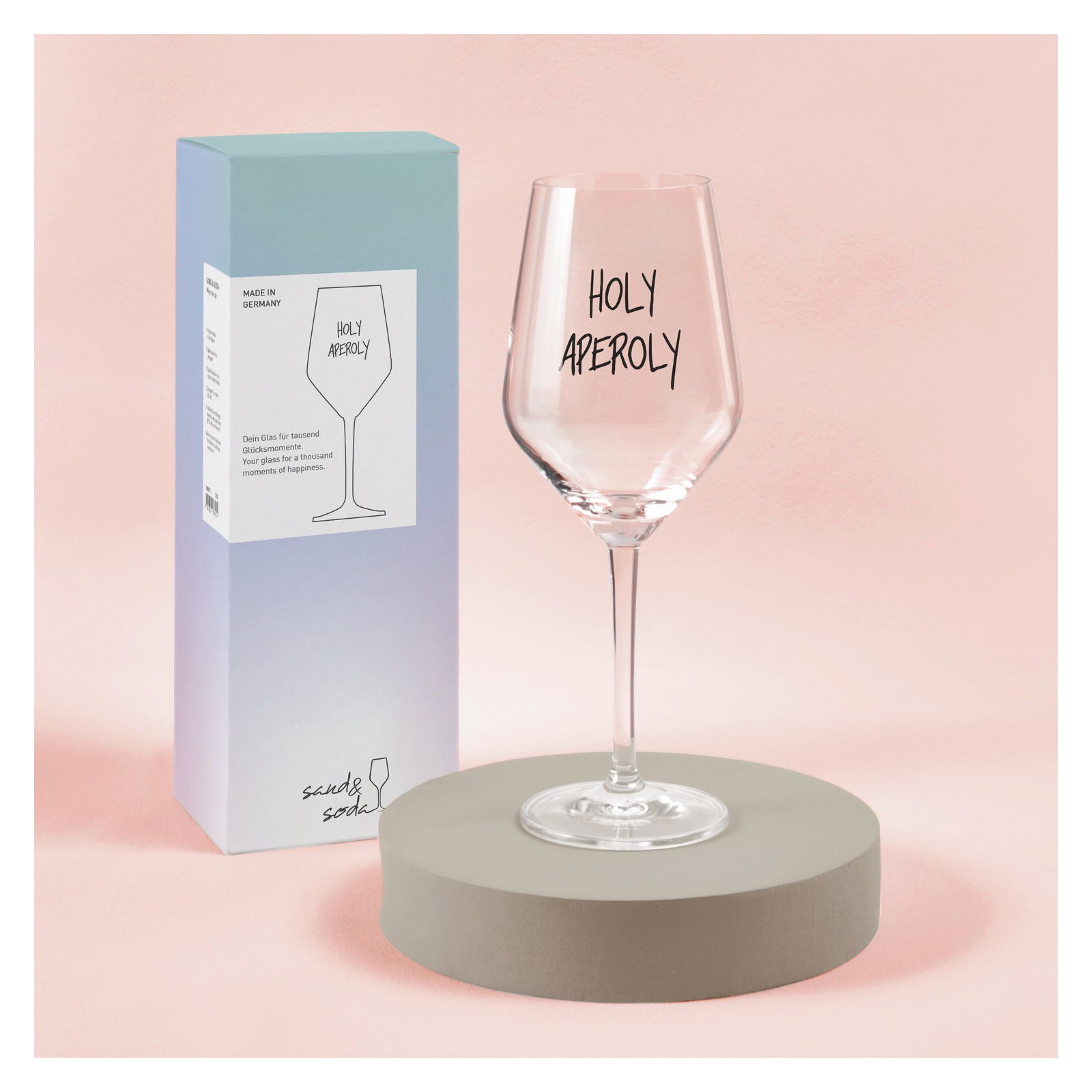 Ritzenhoff Weinglas Holy Aperoly SAND & SODA Weinglas mit der Aufschrift 'Holy Aperoly', präsentiert auf einem runden Podest vor einer Verpackung, aus seitlicher Perspektive.