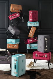 Stilvolle Kofferhocker in verschiedenen Farben, gestapelt in einem eleganten Raum mit dunkler Wandverkleidung, aus seitlicher Perspektive.