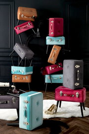 Stilvolle Kofferhocker in verschiedenen Farben, gestapelt in einem eleganten Raum mit dunkler Wandverkleidung, aus seitlicher Perspektive.