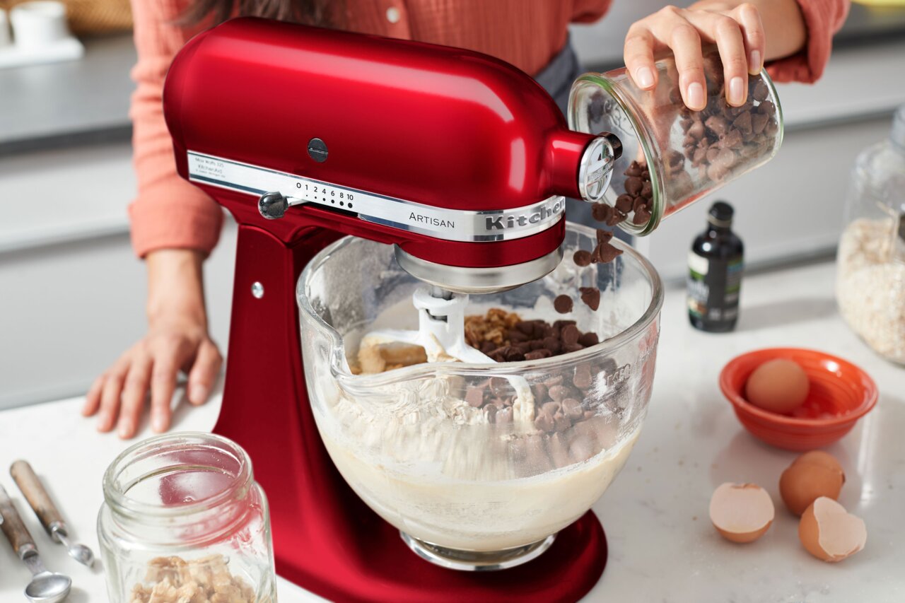 KitchenAid Keukenmachine Liebesapfel ARTISAN Rode keukenmachine 'Liebesapfel' in zijaanzicht, terwijl er chocoladestukjes in de mengkom worden gedaan.