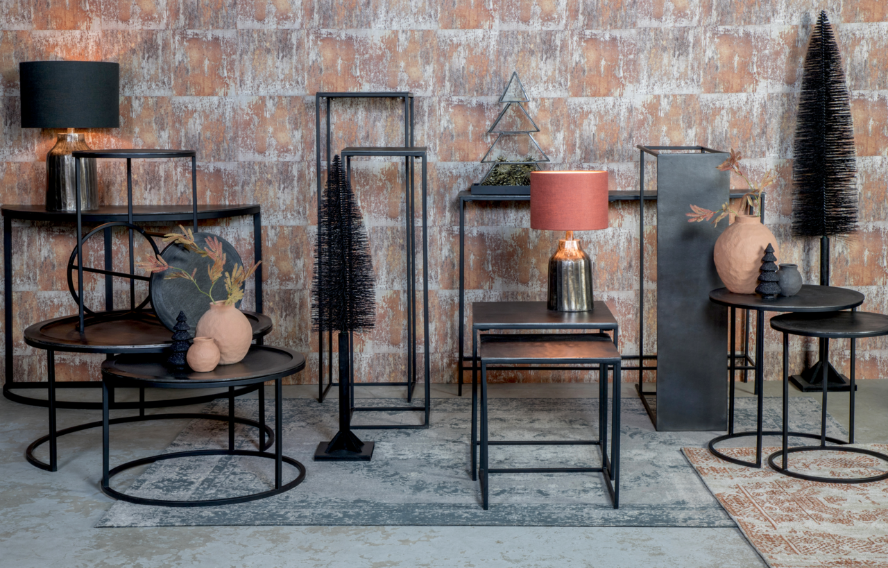 Verschiedene Beistelltische in einem industriellen Design, aus schwarzem Metall mit runden und quadratischen Tischplatten, dekoriert mit Lampen und Vasen, vor einer gemusterten Wand. Perspektive: Frontalansicht.