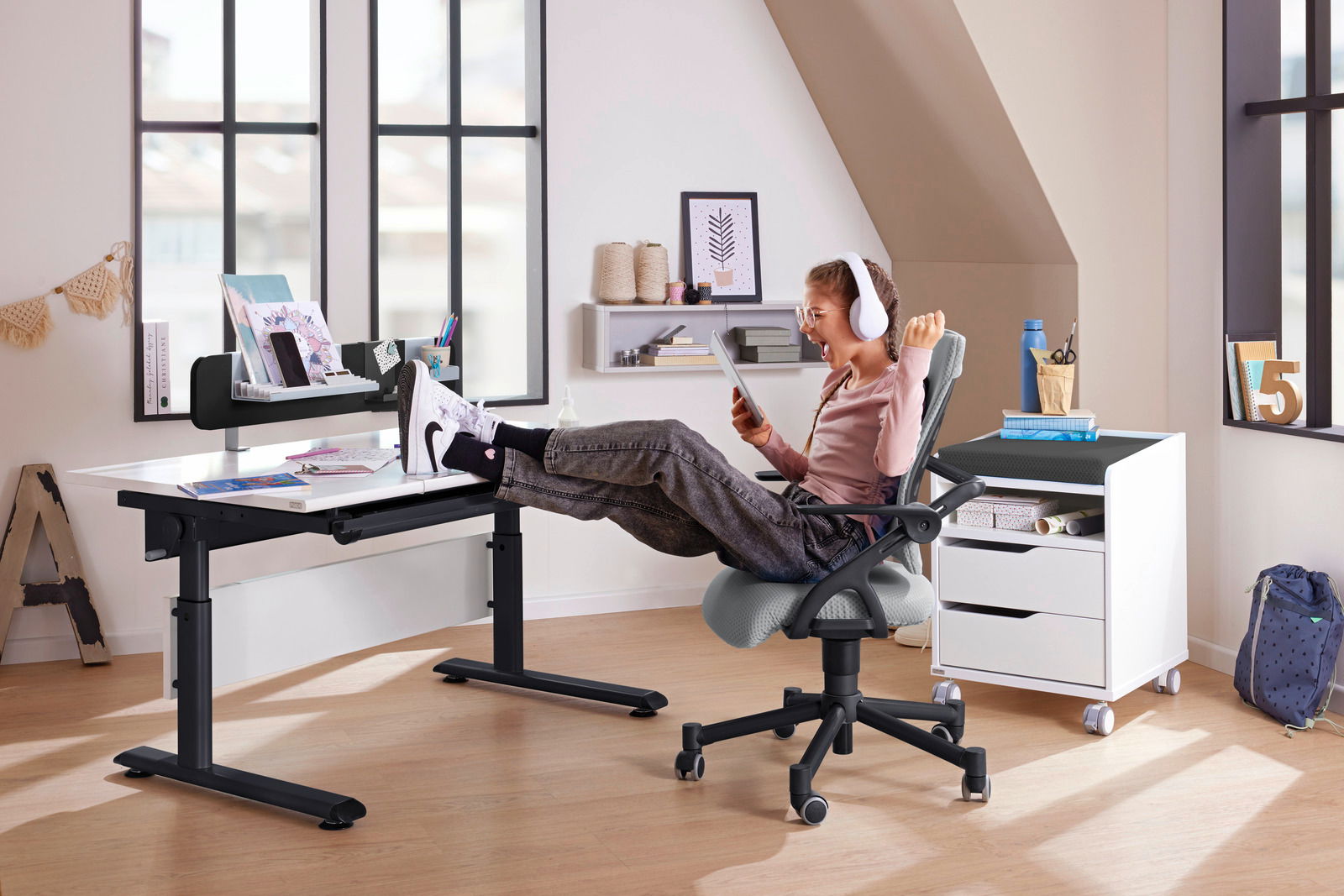 Paidi Schreibtisch MARCO2 Moderner Schreibtisch in einem hellen Raum, mit einem Kind, das entspannt auf einem Bürostuhl sitzt und ein Tablet benutzt. Perspektive von der Seite.