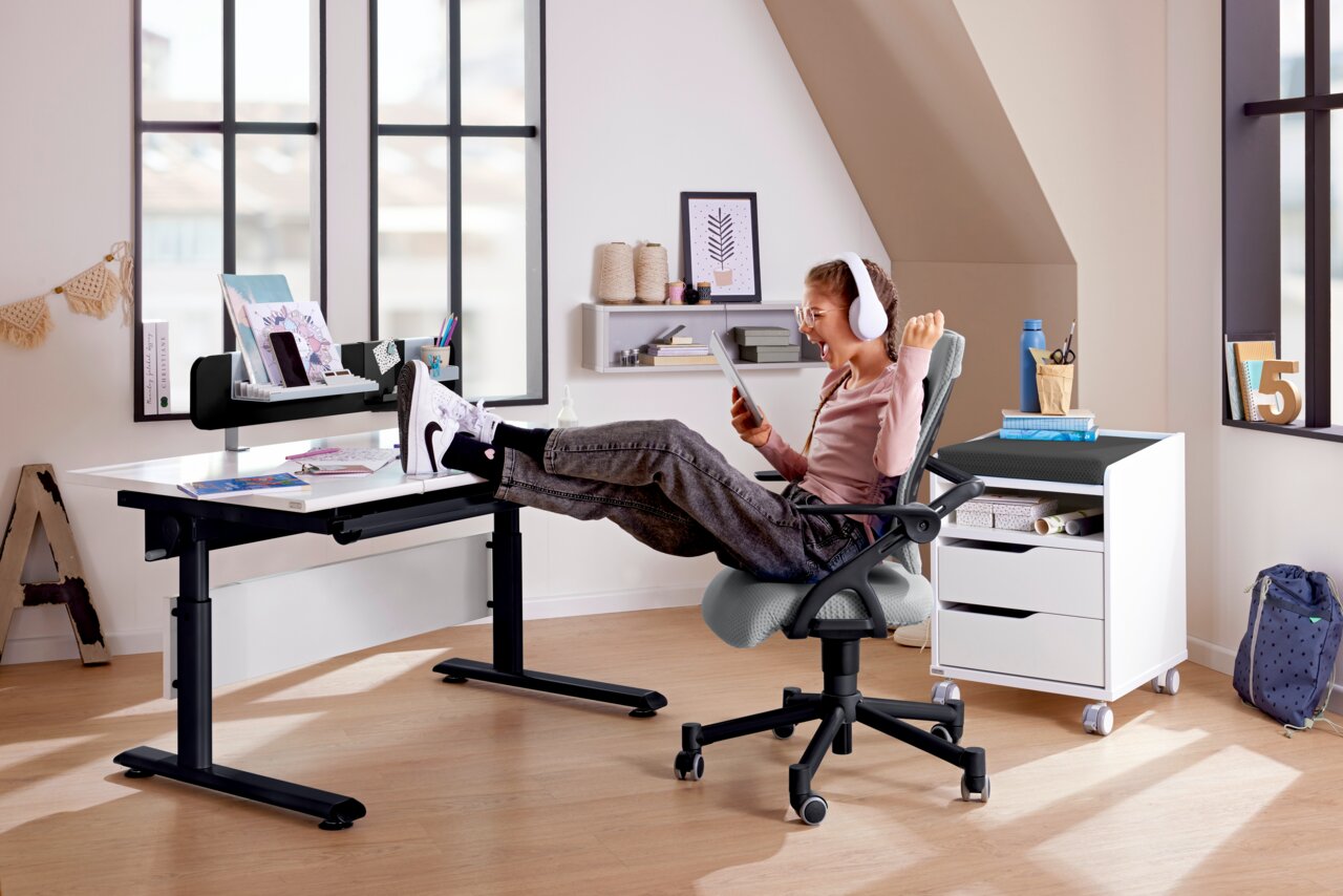 Paidi Schreibtisch MARCO2 Moderner Schreibtisch in einem hellen Raum, mit einem Kind, das entspannt auf einem Bürostuhl sitzt und ein Tablet benutzt. Perspektive von der Seite.