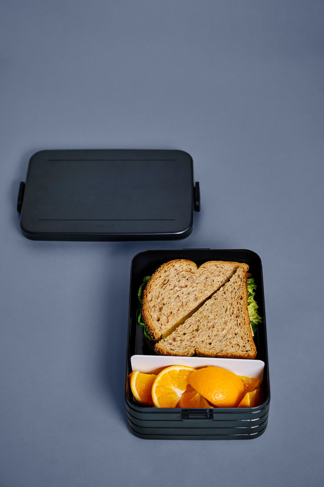 Lunchbox Take a Break Midi in schwarzer Farbe, geöffnet mit einem Sandwich und Orangenscheiben, aus der Vogelperspektive fotografiert.