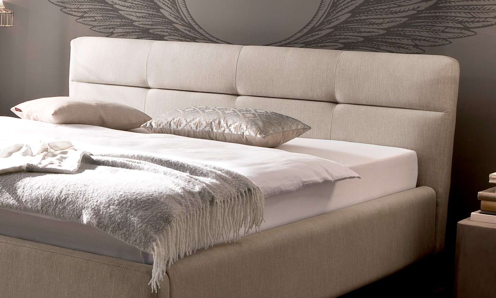 Beiges Polsterbett mit Kopfteil, seitliche Perspektive, dekoriert mit Kissen und Decke.
