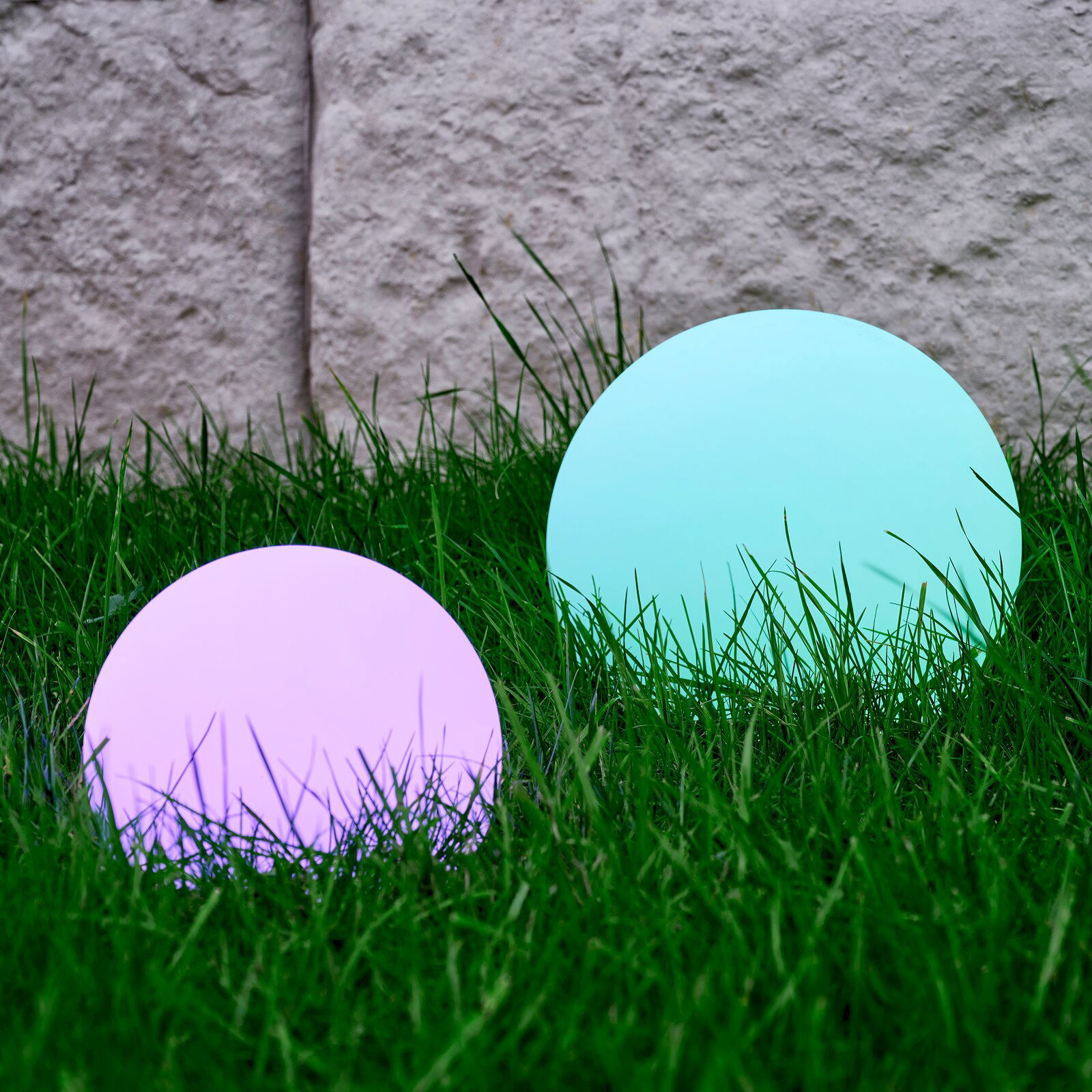 Zwei kugelförmige Solarleuchten von Ambia Garden im Gras, eine in Lila und eine in Türkis, aus seitlicher Perspektive fotografiert.