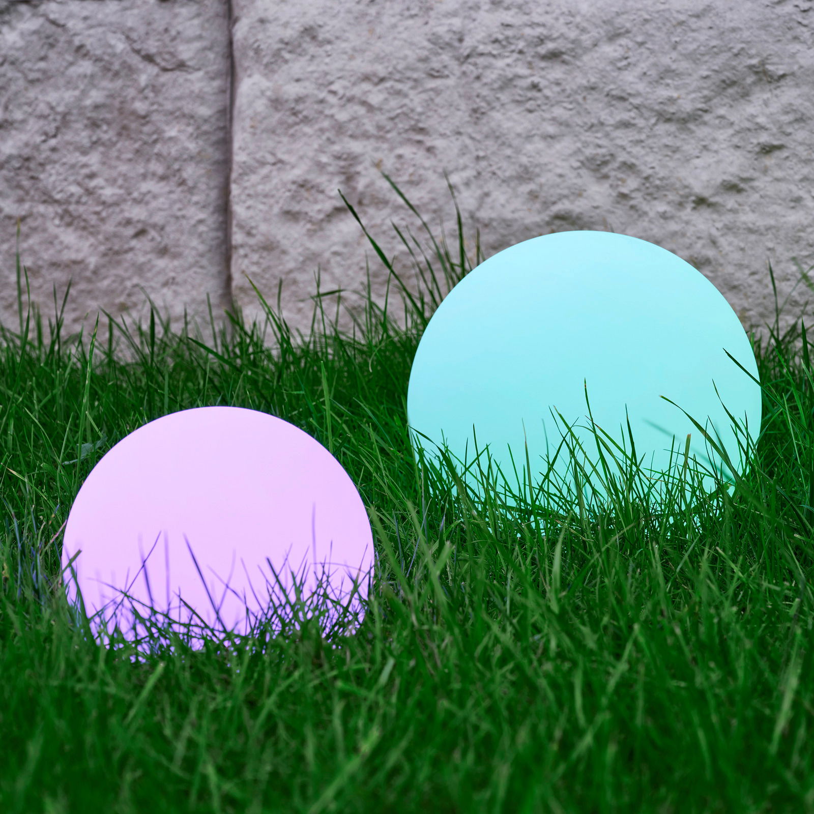 Zwei kugelförmige Solarleuchten von Ambia Garden im Gras, eine in Lila und eine in Türkis, aus seitlicher Perspektive fotografiert.