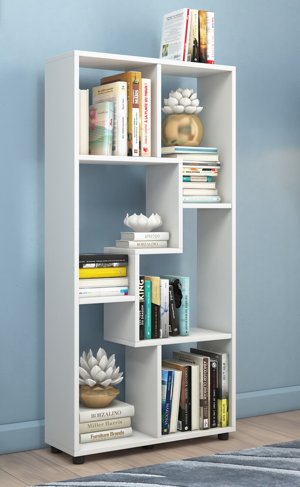 Weißes, modernes Bücherregal mit asymmetrischen Fächern, gefüllt mit Büchern und Dekoration, freistehend vor blauer Wand, Frontansicht leicht schräg von vorn.