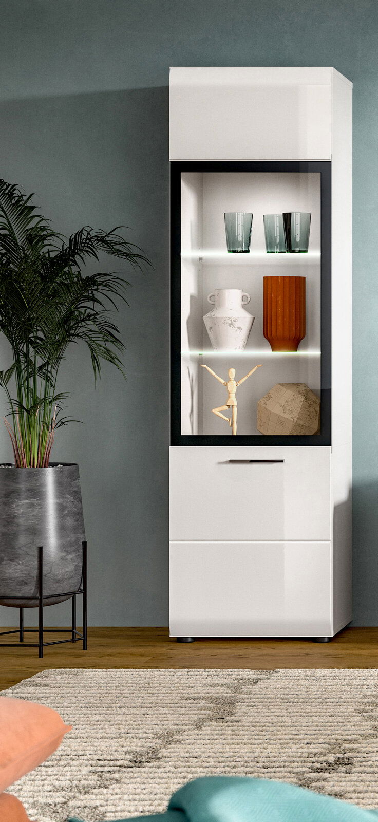 MID.YOU Vitrine ICE Weiße, moderne Vitrine mit schwarzem Rahmen und Glasfront, innen beleuchtet und mit Deko-Objekten bestückt, im Wohnzimmer neben einer großen Topfpflanze; frontale Ansicht mit leichter Perspektive von links.