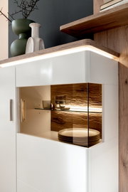 Modernes weißes Sideboard mit beleuchtetem Glasvitrinenfach, Holzrückwand und Metallgriffen; perspektivische Nahaufnahme von schräg vorne rechts