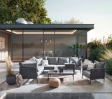 Moderner Loungetisch auf einer Terrasse, umgeben von stilvollen grauen Sofas und Sesseln, mit Blick auf einen Garten und ein modernes Gebäude im Hintergrund.