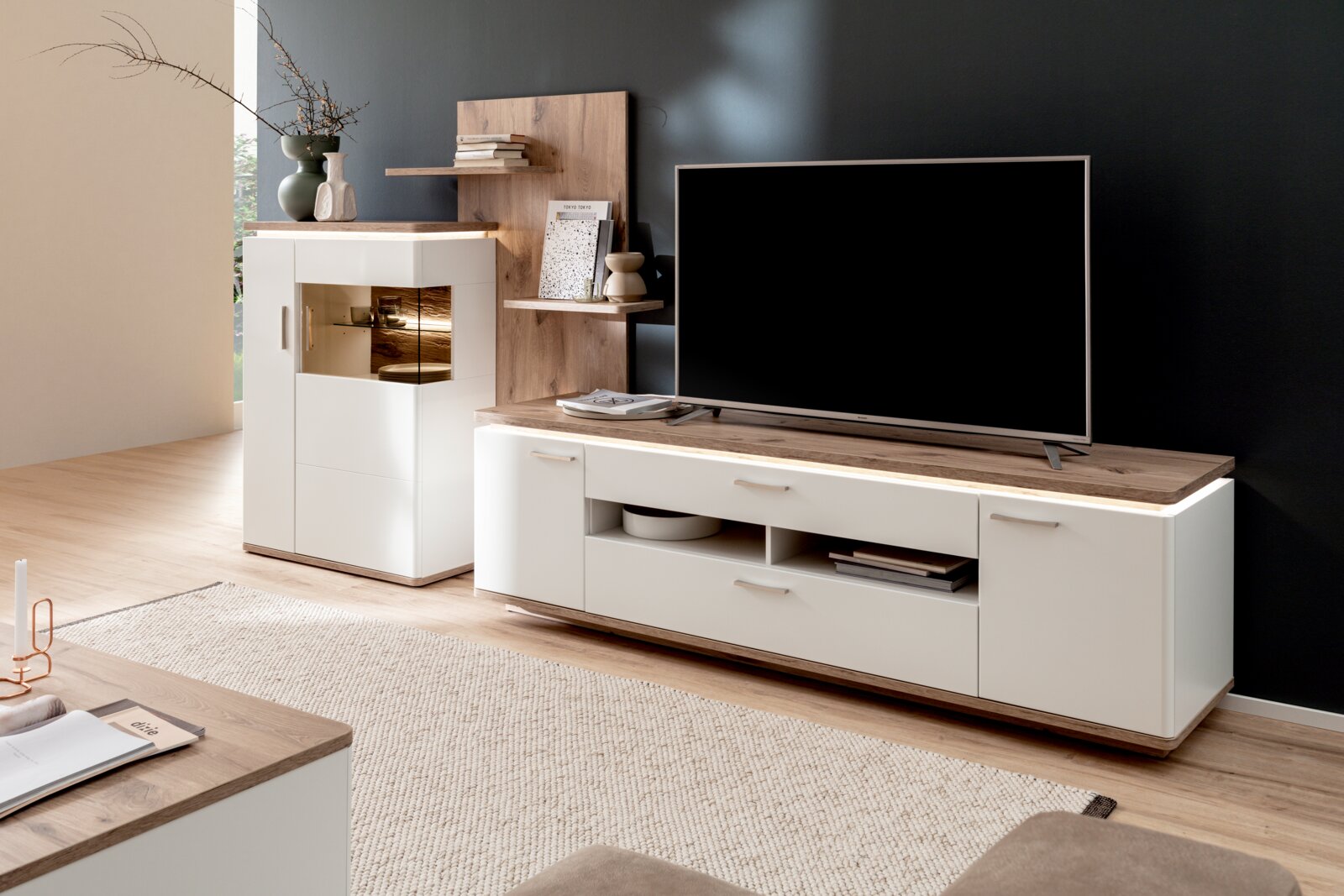 Stylife Wandkombination PALERMO Modernes Wohnzimmer-Set mit weißem TV-Lowboard und Highboard mit Holzoptik-Abdeckplatten und Glasvitrine, dekoriert und mit Fernseher, aufgenommen in leichter Schrägansicht von vorne rechts.