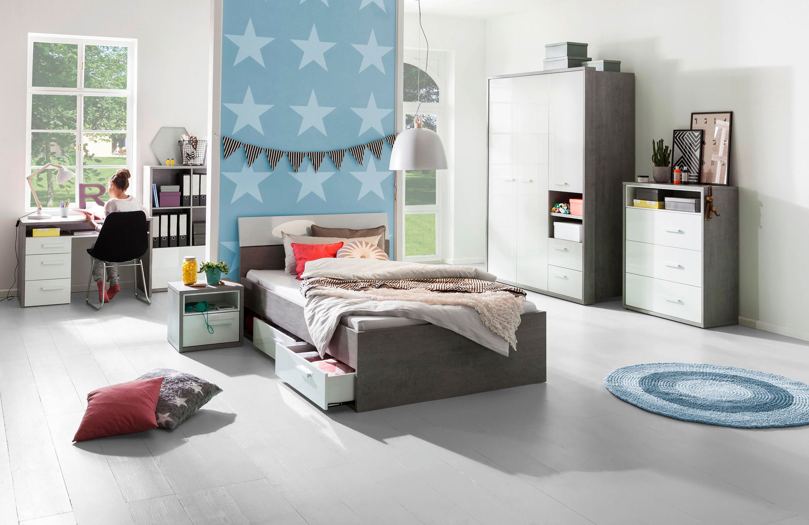 XORA Schreibtisch MARIO Moderne Schlafzimmermöbel mit Stauraumbett, Schreibtisch und Kleiderschrank in einem hellen Raum mit blauer Sternentapete, seitliche Perspektive.