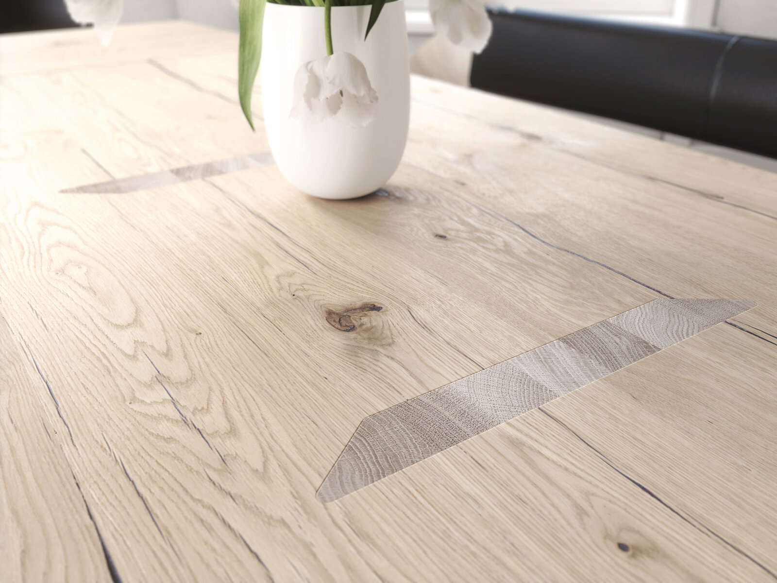 Nahaufnahme eines Voglauer Esstisches V-ROCK aus hellem Holz mit natürlicher Maserung und dekorativen Einlagen, darauf steht eine weiße Vase mit Blumen. Perspektive von schräg oben.