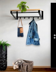 Wandgarderobe mit schwarzem Metallrahmen und Holzablage, darunter mehrere Haken, mit aufgehängter Tasche und Jeansjacke; Perspektive: frontale Ansicht leicht von links