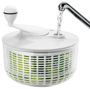 Weiße Salatschleuder mit transparentem Korb und grünen Salatblättern, Wasser wird von oben in die Einfüllöffnung gegossen; Perspektive: Nahaufnahme in leichter Seitenansicht von oben.