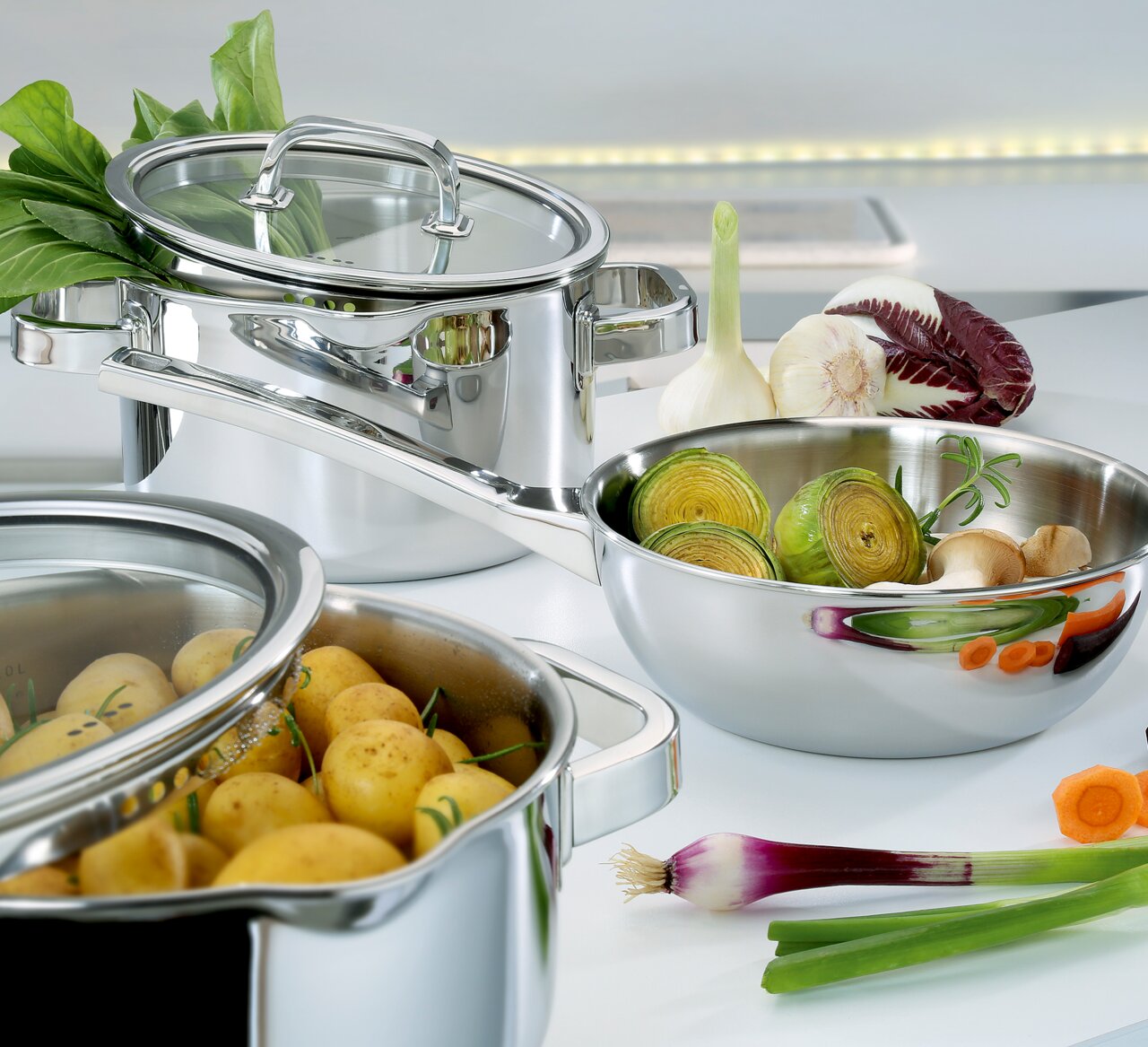 5-delige pannenset FINESSE2+ van roestvrij staal, gevuld met verse groenten en aardappelen, in een moderne keukenomgeving, zijaanzicht
