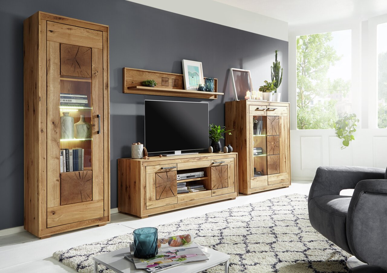 Landscape Wandkombination MERAN Wohnzimmeransicht mit der Landscape Wandkombination MERAN in natürlichem Holzdesign, bestehend aus einem hohen Schrank, einem TV-Board und einem Vitrinenschrank, mit dekorativen Elementen und Pflanzen, seitliche Perspektive.