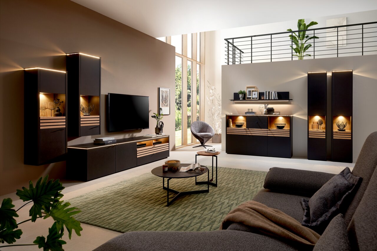 Moderne Wohnzimmer-Wandkombination in dunklen Tönen mit integrierter Beleuchtung, bestehend aus Hängeschränken, TV-Board und Vitrine, aus der Perspektive eines gemütlichen Wohnzimmers mit Sofa und Teppich.