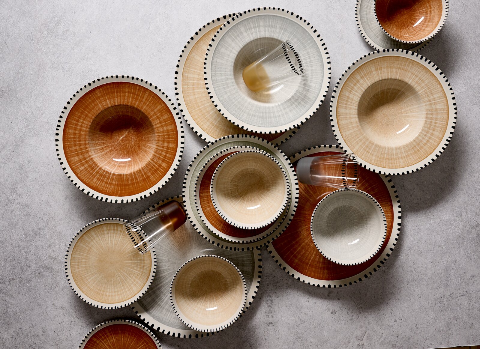Bovenaanzicht van een stijlvol servies met borden, kommen en glazen in warme aardetinten en grijs, afgewerkt met verfijnde zwarte streepjes langs de randen.