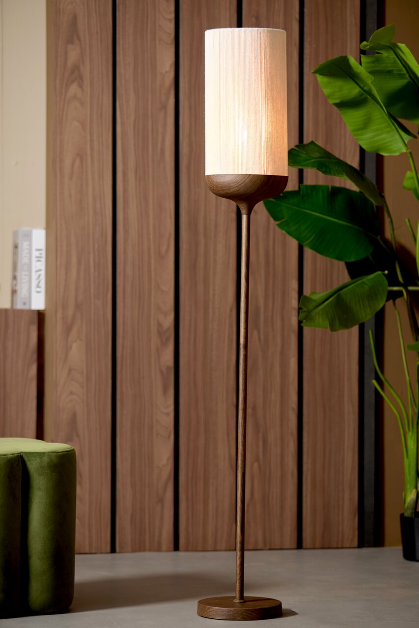 Moderne Stehlampe mit schlankem Holzfuß und zylindrischem, warm leuchtendem Stoffschirm vor Holzpaneelwand, frontale Perspektive.