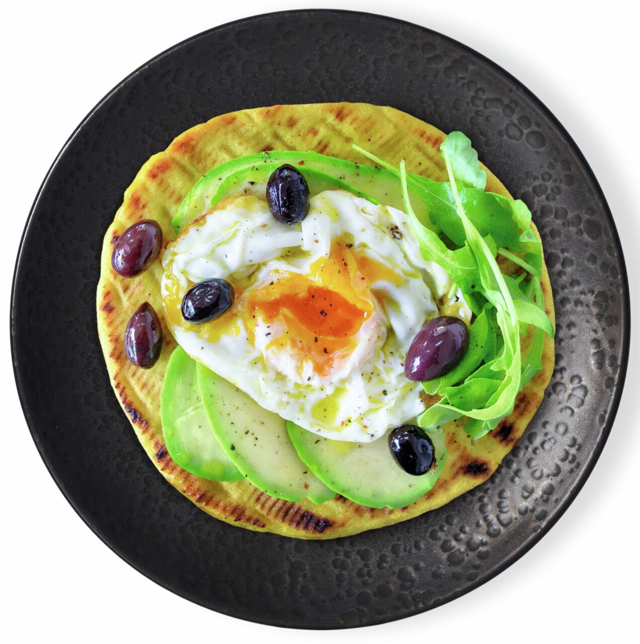 Vogelperspectief op een zwart bord met een belegd flatbread, gegarneerd met avocado, een gepocheerd ei, olijven en rucola.