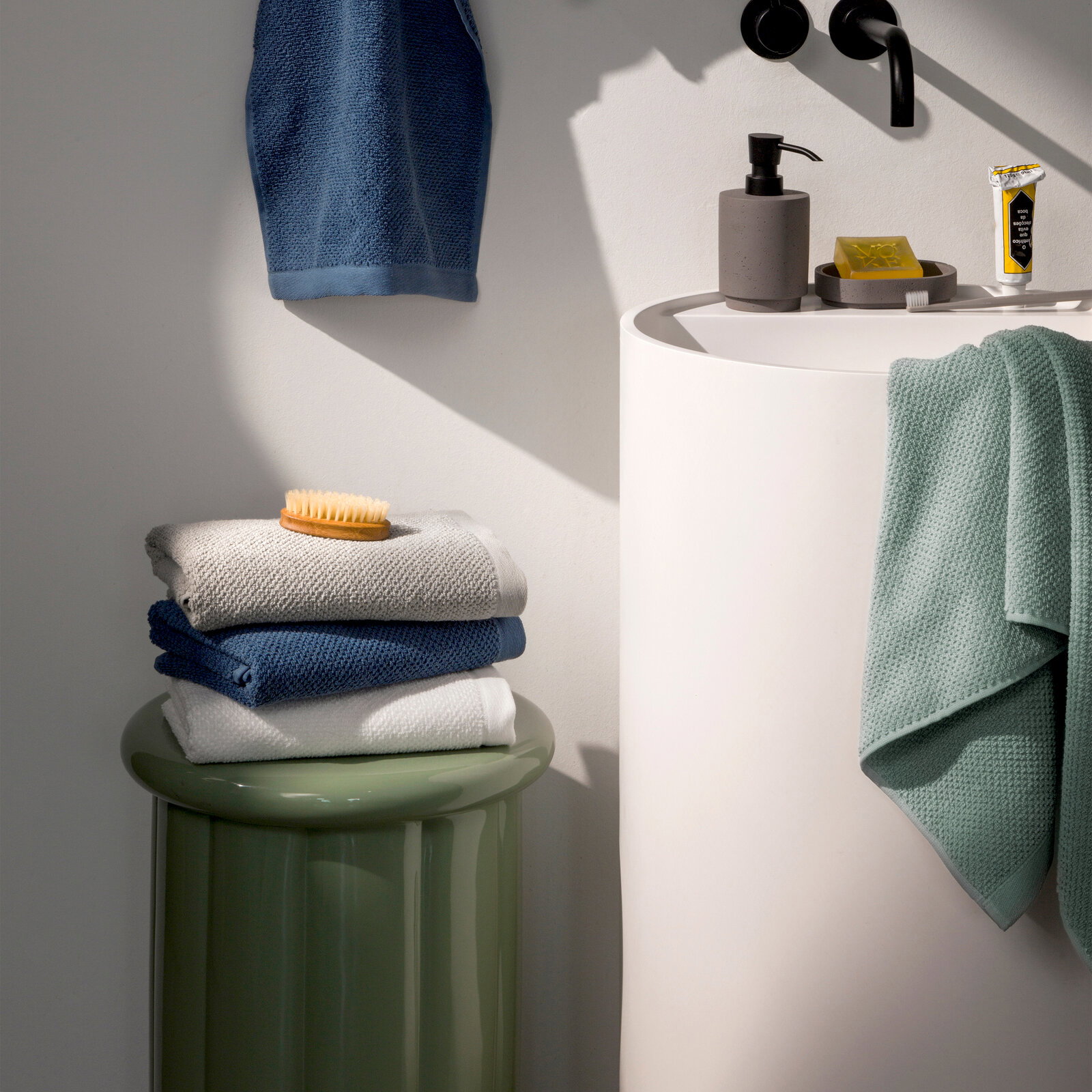 Stapel von Handtüchern in verschiedenen Farben auf einem grünen Hocker neben einem Waschbecken, seitliche Perspektive