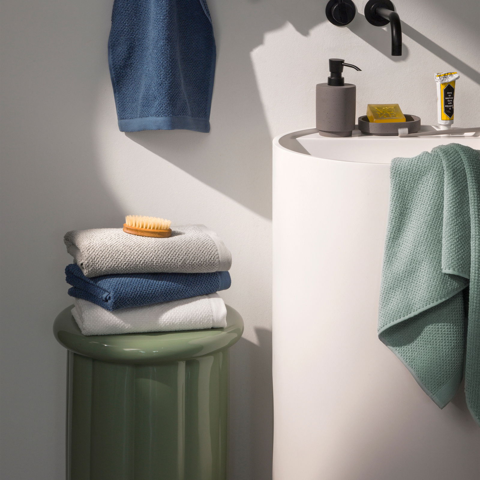 Stapel von Handtüchern in verschiedenen Farben auf einem grünen Hocker neben einem Waschbecken, seitliche Perspektive