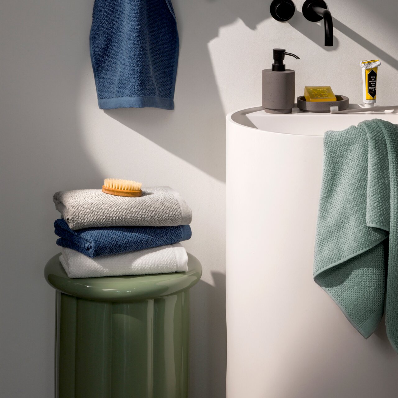 MÖVE Handtuchset 10-tlg. BLISS Stapel von Handtüchern in verschiedenen Farben auf einem grünen Hocker neben einem Waschbecken, seitliche Perspektive