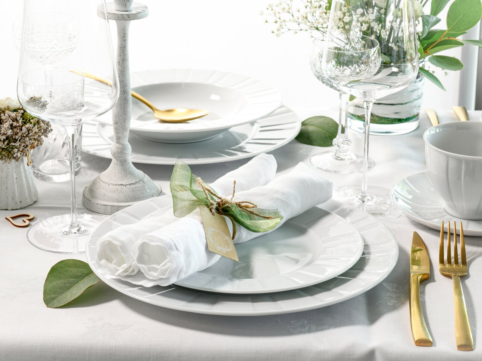 Elegant gedeckter Tisch mit weißen Porzellantellern, gerollter Serviette mit grüner Schleife und Namenskarte, Kristallgläsern und goldfarbenem Besteck, seitliche Perspektive mit leicht erhöhter Ansicht.
