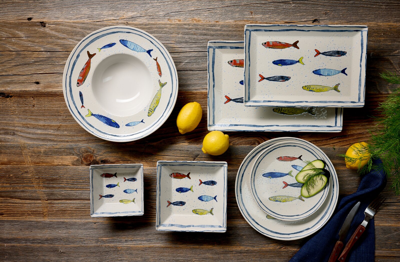 Draufsicht auf rustikales weißes Keramikgeschirr mit handgemalten bunten Fischmotiven, arrangiert mit Zitronen und Gurkenscheiben auf einem Holztisch