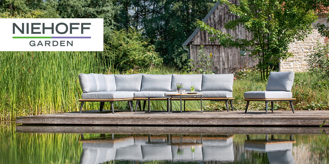 Ein Sitzbereich im Freien mit grauen Kissen, einem Holztisch und einer Teichkulisse, Design von Niehoff Garden.