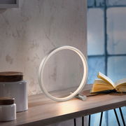 Moderne Tischleuchte MINI RITUS in Ringform auf einem Tisch, seitliche Perspektive, neben einem geöffneten Buch.