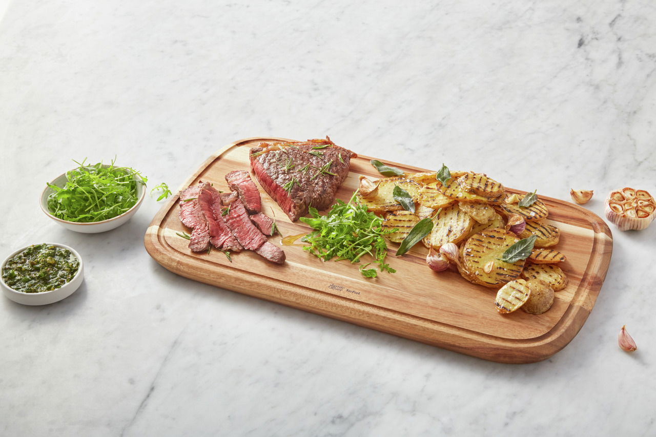 Großes Schneidebrett aus Holz mit gegrilltem Fleisch, Kartoffelscheiben und frischen Kräutern, von oben fotografiert.