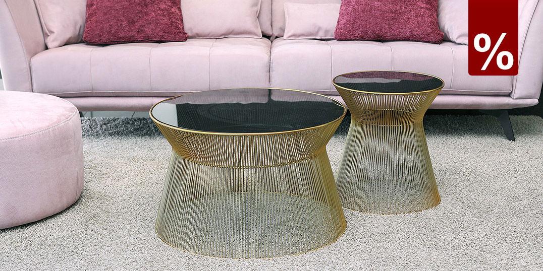 Wohnzimmer mit rosafarbenem Sofa, violetten Kissen und zwei goldenen Draht-Couchtischen mit dunkler Glasplatte auf hellem Teppich