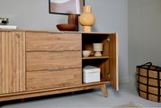 Holz-Sideboard mit drei Schubladen und einer offenen Tür, die dekorative Gegenstände zeigt. Perspektive von vorne links.