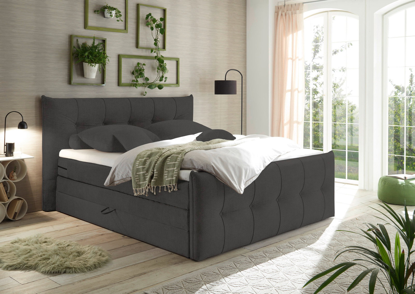 Boxspringbett in einem modernen Schlafzimmer, seitliche Perspektive, mit grauem Stoffbezug, dekoriert mit Pflanzen und einem großen Fenster im Hintergrund.