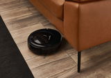 Ein schwarzer Saugroboter reinigt den Holzboden unter einem braunen Ledersofa aus der Perspektive von schräg oben.