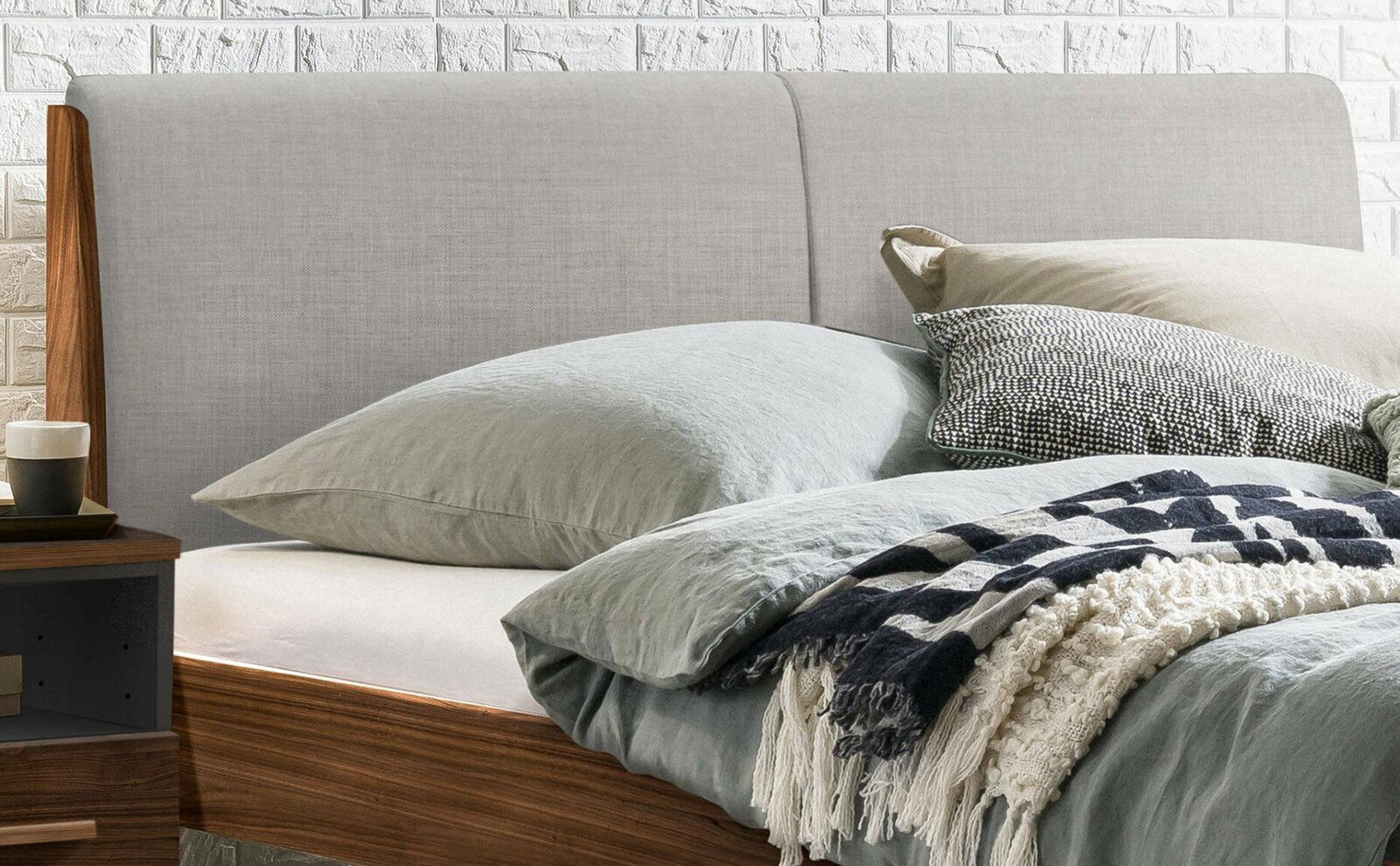 Nahaufnahme eines Bettgestells mit grauem Kopfteil, dekoriert mit Kissen und Decken, seitliche Perspektive