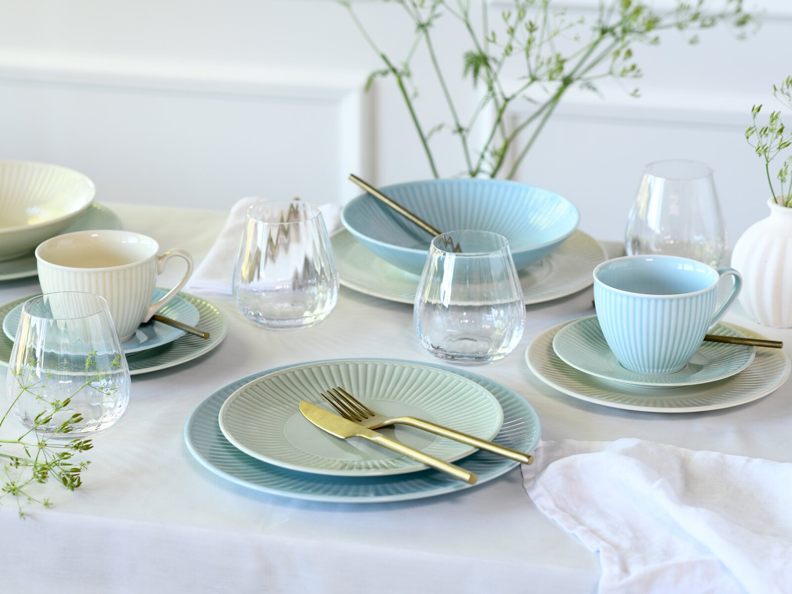 Pastellfarbenes Geschirr-Set in Hellblau und Creme mit gerippten Tellern, Schalen und Tassen, dazu klare Wassergläser und goldfarbenes Besteck auf weißer Tischdecke, fotografiert aus leicht erhöhter schräger Perspektive von vorn.