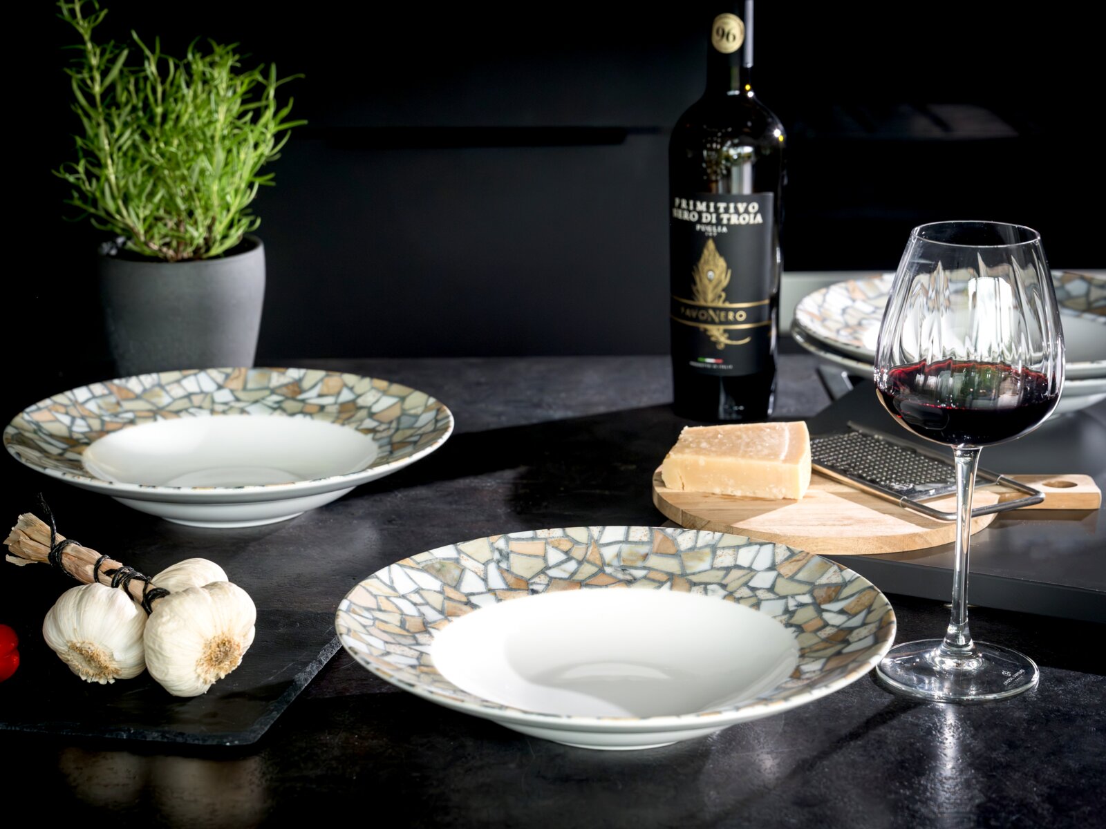 Modernes Tisch-Arrangement mit zwei tiefen Keramikschalen im Mosaik-Design, Rotweinglas, Weinflasche, Parmesan auf Holzbrett mit Reibe, Knoblauch und Kräutertopf auf dunkler Arbeitsplatte, fotografiert aus leicht erhöhter Frontperspektive.