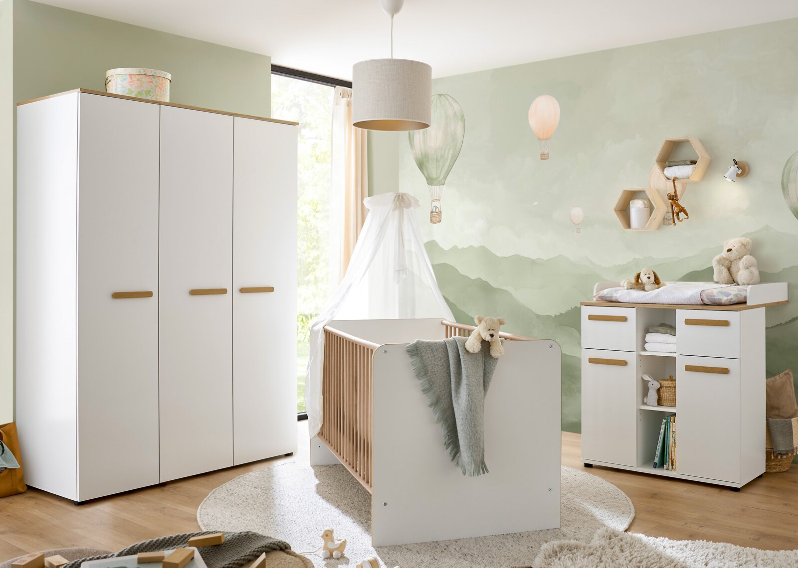 MyBaby Kleiderschrank ASTEA Helles Babyzimmer mit weißem Babybett mit Holzstäben, großem weißen Kleiderschrank und Wickelkommode mit Holzgriffen vor Wandmotiv mit Heißluftballons, frontal aus leicht seitlicher Perspektive aufgenommen.