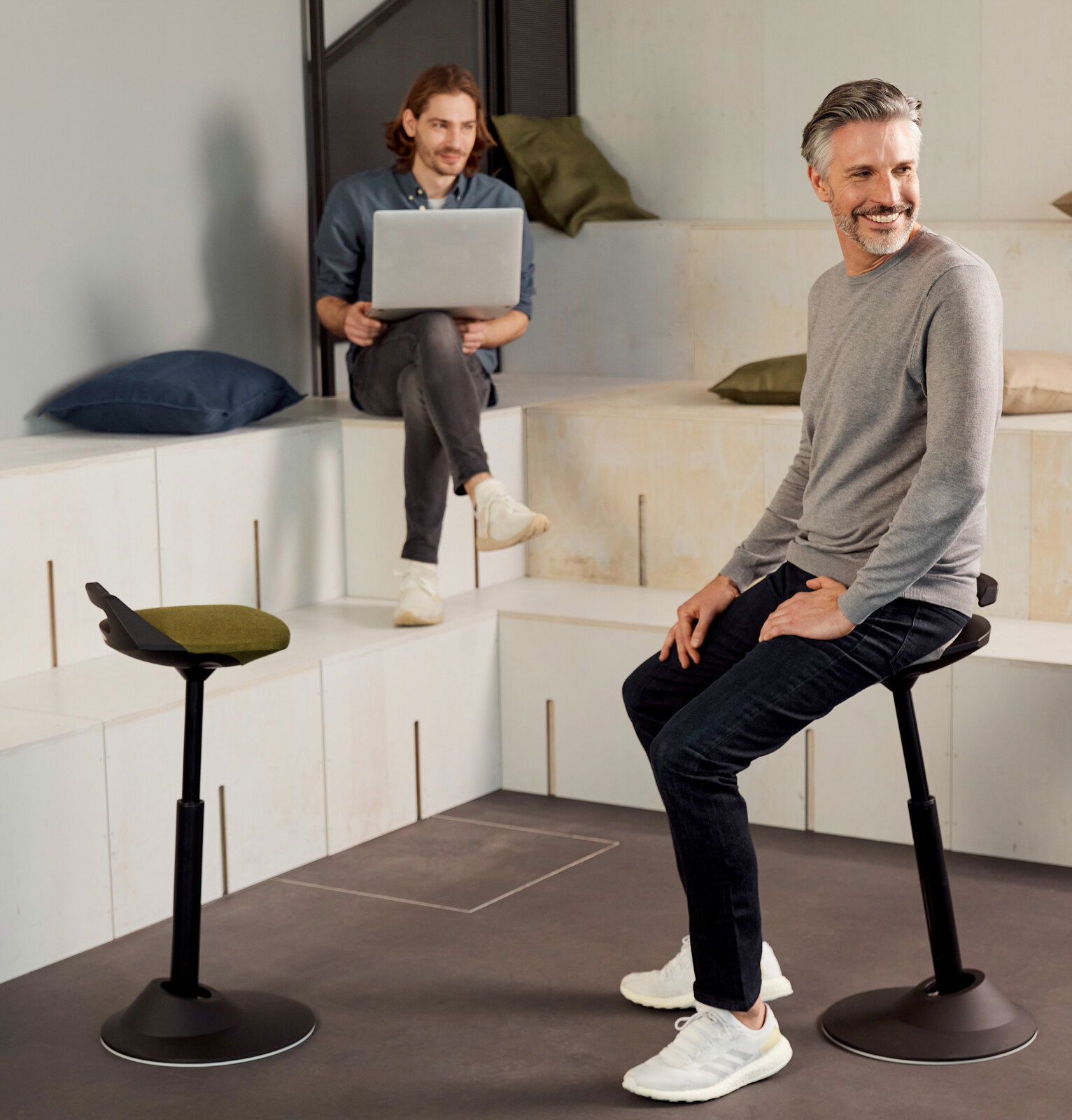 Zwei Männer in einem modernen Raum, einer sitzt auf einer Aeris Sitz-/Stehhilfe Muvman, seitliche Perspektive.