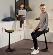 Zwei Männer in einem modernen Raum, einer sitzt auf einer Aeris Sitz-/Stehhilfe Muvman, seitliche Perspektive.