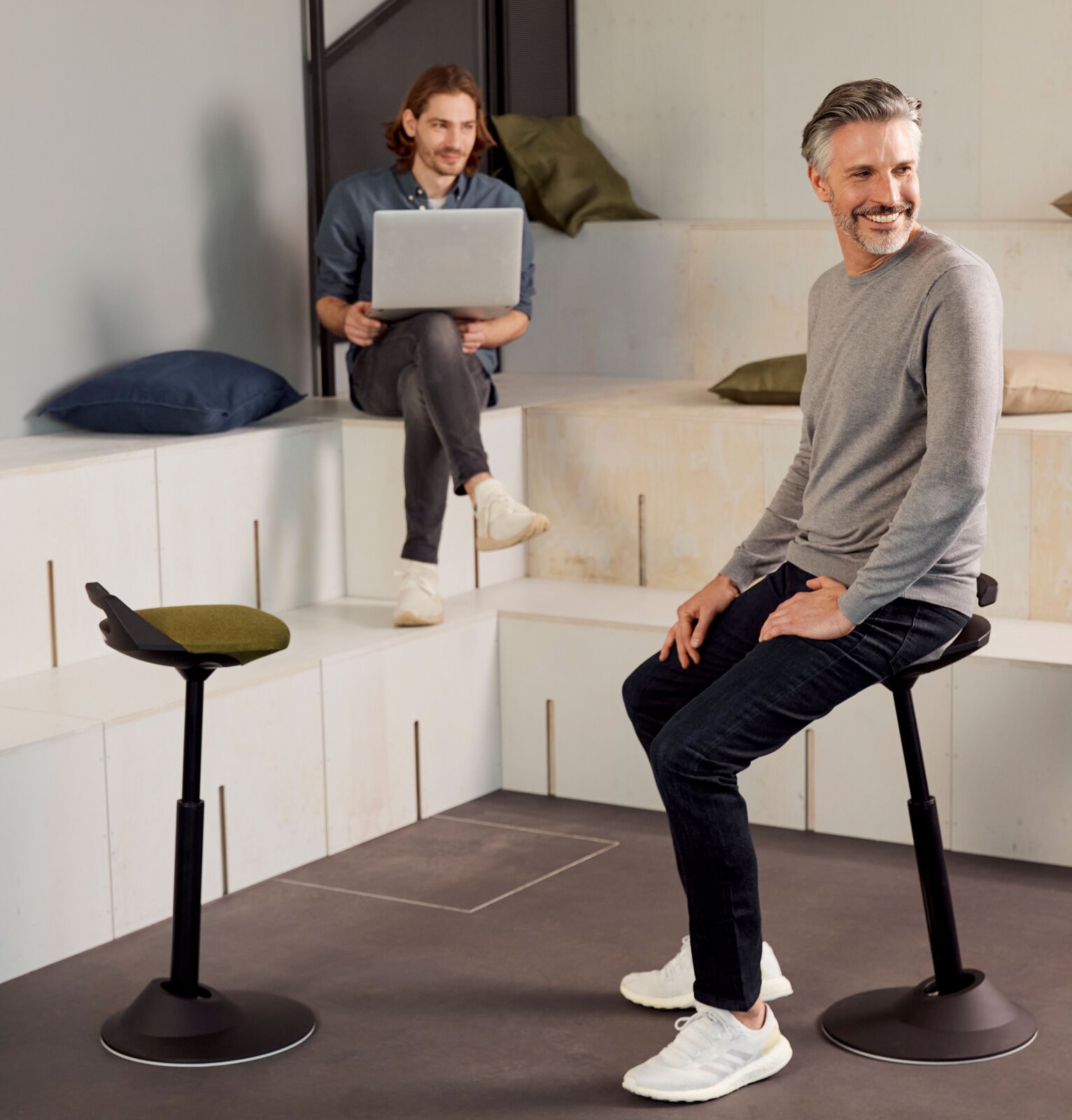 Zwei Männer in einem modernen Raum, einer sitzt auf einer Aeris Sitz-/Stehhilfe Muvman, seitliche Perspektive.